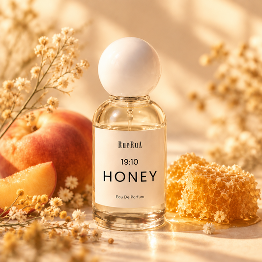 A clear glass perfume bottle with a white spherical cap and a white label reading RueBul 19:10 HONEY Eau De Parfum, set against a light gray background.