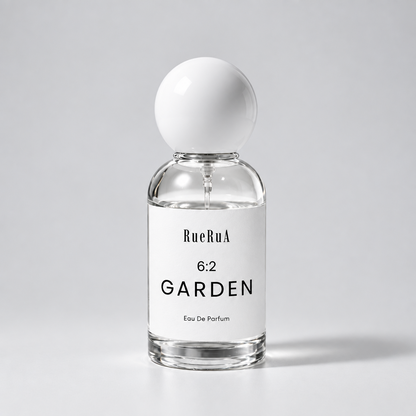 A clear glass perfume bottle with a white spherical cap and a white label reading RueBula 6:2 GARDEN Eau De Parfum, set against a light gray background.