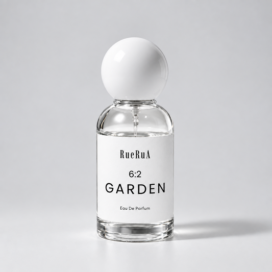 A clear glass perfume bottle with a white spherical cap and a white label reading RueBula 6:2 GARDEN Eau De Parfum, set against a light gray background.